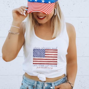 May include: White tank top featuring an American flag design with the text "stars stripes and LATE STAGE CAPITALISM fuck donald trump". The person in the image is wearing a red, white, and blue baseball cap.