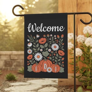 Può includere: Bandiera da giardino nera con la scritta "Welcome" in corsivo bianco. La bandiera presenta un disegno floreale con zucche arancioni, margherite e papaveri. Le lettere "FDT" sono sulle zucche. La bandiera è esposta su un supporto in metallo nero.