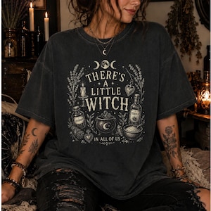 Puede incluir: Una camiseta gris carbón de manga corta con un estampado gráfico en color crema que incluye el texto "THERE'S A LITTLE WITCH IN ALL OF US." El diseño presenta ilustraciones de una vela, una botella, un caldero y elementos botánicos.