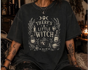 Könnte beinhalten: Ein anthrazitfarbenes Kurzarm-T-Shirt mit einem cremefarbenen Grafikdruck und dem Text "THERE'S A LITTLE WITCH IN ALL OF US." Das Design zeigt Illustrationen einer Kerze, einer Flasche, eines Kessels sowie verschiedene botanische Elemente.