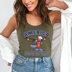 May include: Olive green tank top with the word "AMERICA" in blue and white lettering. A cartoon hot dog character is grilling on a barbecue. The text "GRILLING THROUGH THE EXISTENTIAL DREAD" is below the graphic.