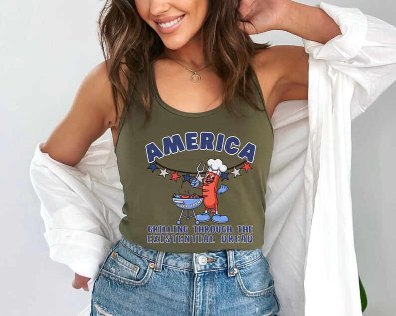 May include: Olive green tank top with the word "AMERICA" in blue and white lettering. A cartoon hot dog character is grilling on a barbecue. The text "GRILLING THROUGH THE EXISTENTIAL DREAD" is below the graphic.