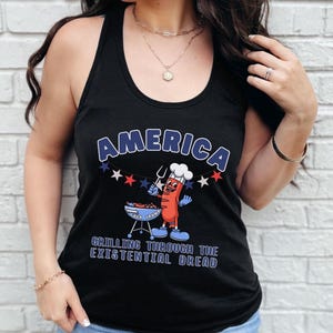 May include: Black tank top with the word "AMERICA" in blue and white lettering. A cartoon hot dog character is grilling on a barbecue. The text "GRILLING THROUGH THE EXISTENTIAL DREAD" is below the graphic.
