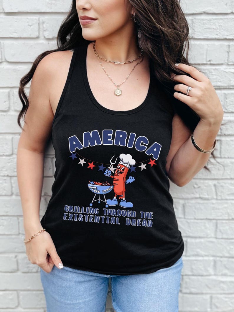 May include: Black tank top with the word "AMERICA" in blue and white lettering. A cartoon hot dog character is grilling on a barbecue. The text "GRILLING THROUGH THE EXISTENTIAL DREAD" is below the graphic.