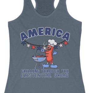 May include: A blue-grey tank top with the word "AMERICA" in a white and blue font. A cartoon hot dog character is grilling on a barbecue. The text "GRILLING THROUGH THE EXISTENTIAL DREAD" is below the cartoon.