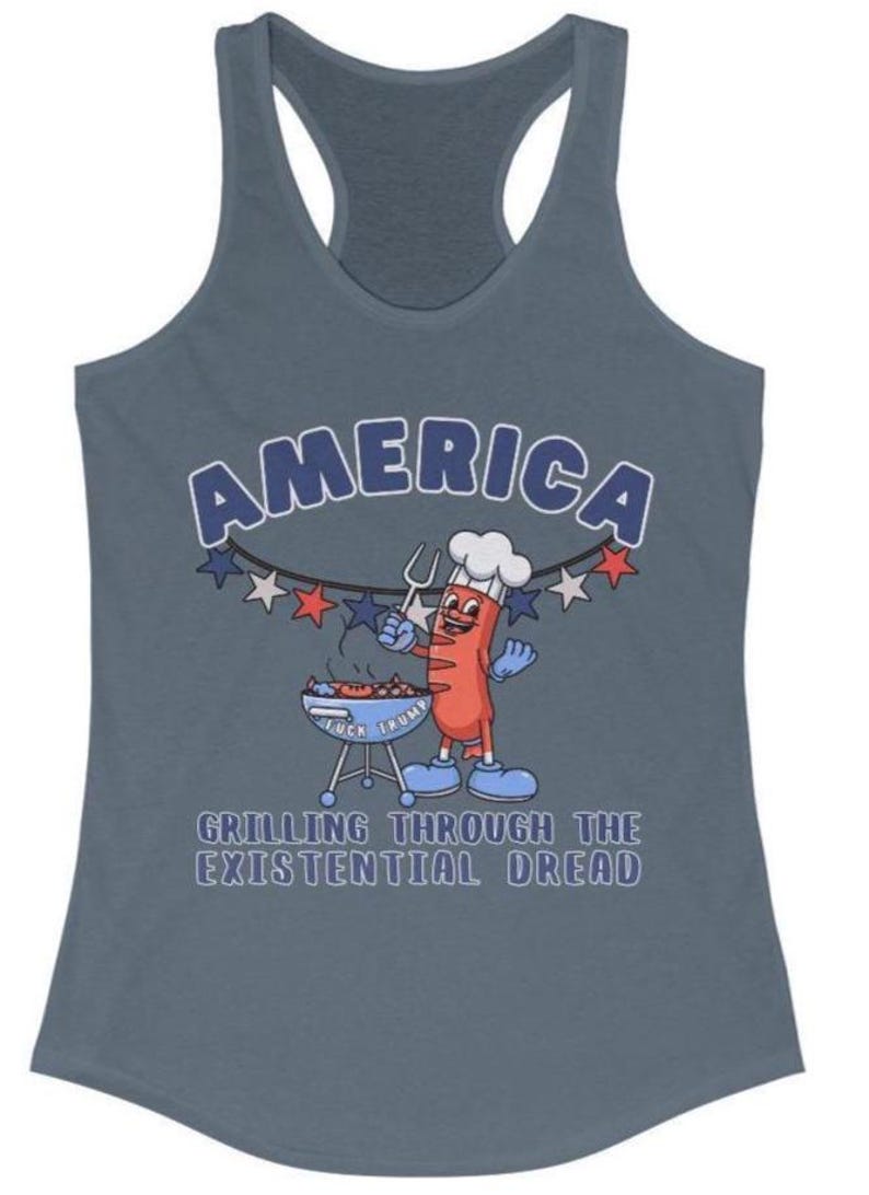May include: A blue-grey tank top with the word "AMERICA" in a white and blue font. A cartoon hot dog character is grilling on a barbecue. The text "GRILLING THROUGH THE EXISTENTIAL DREAD" is below the cartoon.
