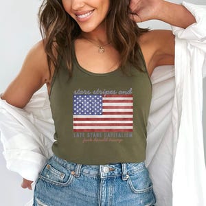 May include: Olive green tank top featuring an American flag design with the text "stars stripes and late stage capitalism fuck donald trump". The flag has red and white stripes and a blue field with white stars.