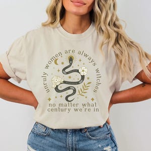 May include: A beige t-shirt with a graphic print of a snake with stars and a crescent moon. The text on the shirt reads "Unruly women are always witches no matter what century we're in."