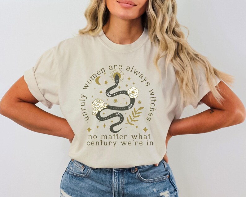 May include: A beige t-shirt with a graphic print of a snake with stars and a crescent moon. The text on the shirt reads "Unruly women are always witches no matter what century we're in."