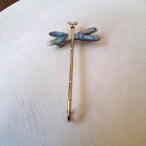 May include: A gold-colored dragonfly hair pin with iridescent blue and green wings. The dragonfly has a long, slender body and a curved hook at the end. The wings are oval-shaped and have a pearlescent sheen.
