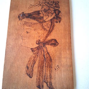May include: A wood-burned portrait of a woman in profile, wearing a hat adorned with flowers and a large bow. The artwork is on a rectangular wooden plaque, with the initials "A.J.T." in the lower right corner. The wood has a warm, natural tone.