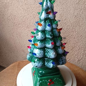 May include: A ceramic Christmas tree with a green base and body, adorned with colorful light bulbs. The tree has a snow-like effect on its branches and is placed on a white surface. The base is decorated with red berries.