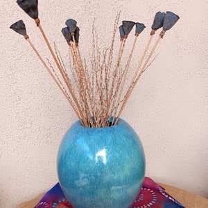 May include: A turquoise blue, round ceramic vase holds dried floral stems and dark blue seed pods on wooden sticks. The vase sits on a colorful, patterned fabric.
