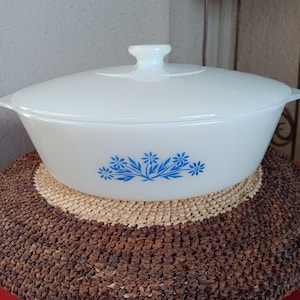 May include: White casserole dish with a blue floral design and a white lid. The dish is sitting on a brown and beige woven mat.