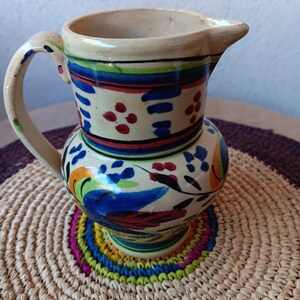 May include: A white ceramic pitcher with a blue, red, and yellow floral design. The pitcher has a handle and a spout.