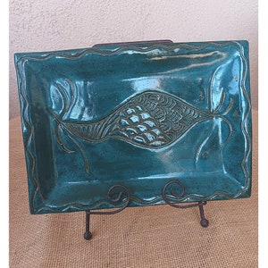 May include: Teal ceramic serving tray with a raised design of two fish with scales. The tray has a wavy edge and is on a black metal stand.