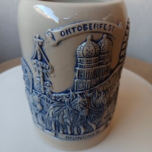 May include: A ceramic beer stein with a blue and white design depicting a scene from the Oktoberfest celebration in Munich, Germany. The stein features a banner with the text "OKTOBERFEST" and "MUNCHEN".