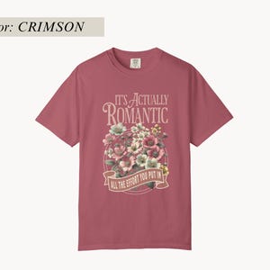 Pu&ograve; includere: T-shirt color cremisi con grafica floreale. La grafica include un bouquet di fiori rosa, bianchi e verdi, con la scritta "IT'S ACTUALLY ROMANTIC" sopra uno striscione con la scritta "ALL THE EFFORT YOU PUT IN."
