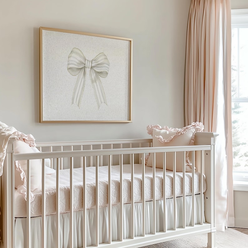 May include: A white cot with a pink and white bedding set. The cot has a white frill around the bottom and a white pillow with a pink frill. A framed print of a white bow with grey stripes hangs on the wall behind the cot.