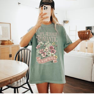 Pu&ograve; includere: T-shirt verde chiaro con la scritta "IT'S ACTUALLY ROMANTIC" sopra un disegno floreale. Il disegno comprende fiori rosa e bianchi. Il testo "ALL THE EFFORT YOU PUT IN" &egrave; in basso. Indossato un berretto da baseball nero.