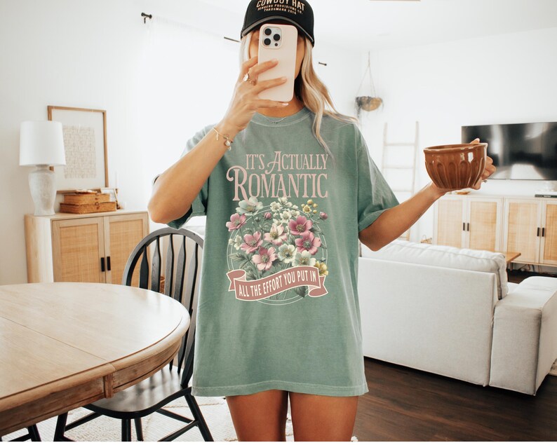 Pu&ograve; includere: T-shirt verde chiaro con la scritta "IT'S ACTUALLY ROMANTIC" sopra un disegno floreale. Il disegno comprende fiori rosa e bianchi. Il testo "ALL THE EFFORT YOU PUT IN" &egrave; in basso. Indossato un berretto da baseball nero.