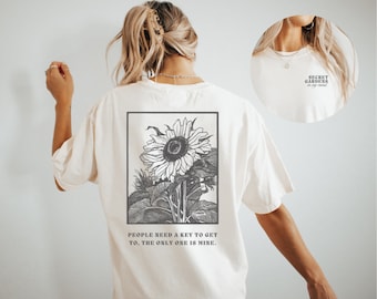 May include: A white t-shirt with a black and white illustration of a sunflower and the text "PEOPLE NEED A KEY TO GET TO, THE ONLY ONE IS MINE." on the back. The front of the shirt has the text "SECRET GARDENS" in black.