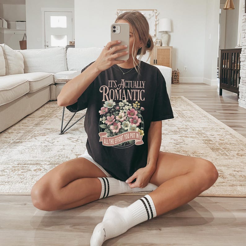 Pu&ograve; includere: T-shirt nero con la scritta "IT'S ACTUALLY ROMANTIC" sopra un disegno floreale. Il disegno floreale comprende fiori rosa, bianchi e verdi. La maglietta ha anche la scritta "ALL THE EFFORT YOU PUT IN" su uno striscione sottostante.