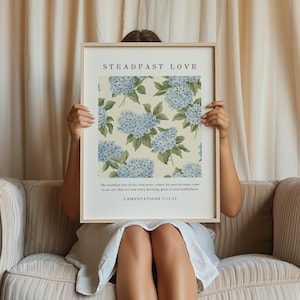 May include: A framed print with a floral design of blue hydrangeas on a cream background. The print has the text "STEADFAST LOVE" at the top and a Bible verse from Lamentations 3:22-23 at the bottom.