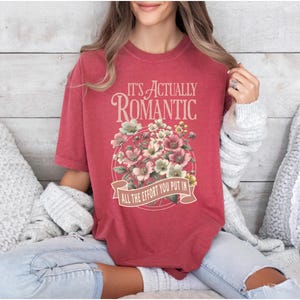Pu&ograve; includere: T-shirt rosso con la scritta "IT'S ACTUALLY ROMANTIC" sopra un disegno floreale. Il disegno comprende fiori rosa e bianchi. Sotto i fiori c'&egrave; uno striscione con la scritta "ALL THE EFFORT YOU PUT IN".