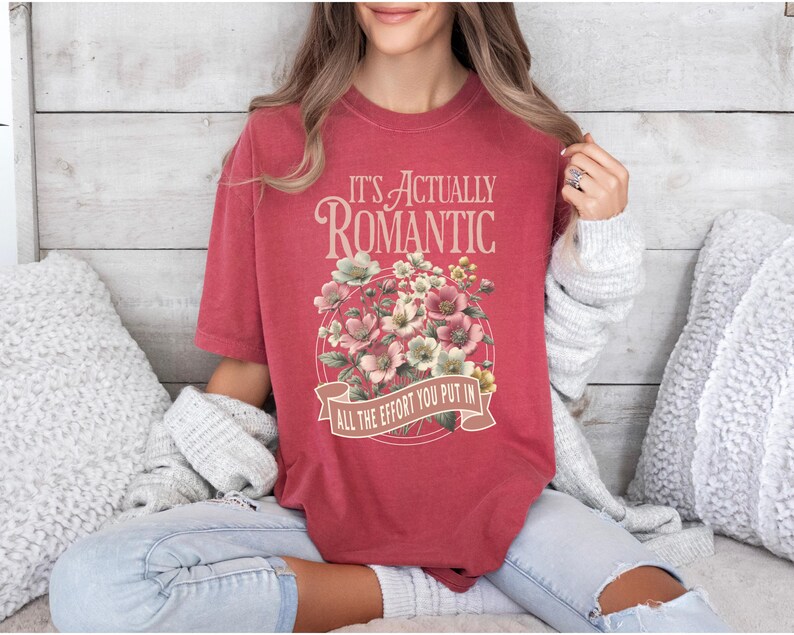 Pu&ograve; includere: T-shirt rosso con la scritta "IT'S ACTUALLY ROMANTIC" sopra un disegno floreale. Il disegno comprende fiori rosa e bianchi. Sotto i fiori c'&egrave; uno striscione con la scritta "ALL THE EFFORT YOU PUT IN".