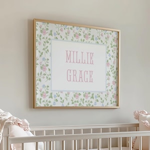 May include: A framed print with the name "MILLIE GRACE" in pink script. The print has a floral pattern with pink and blue flowers on a white background, framed in gold. The print is hanging above a white crib.