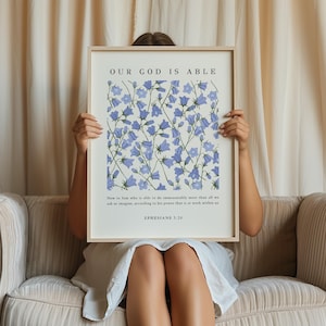 May include: A framed print featuring a floral design and the text "OUR GOD IS ABLE" and a Bible verse from Ephesians 3:20. The print is being held by a person in front of a white curtain.