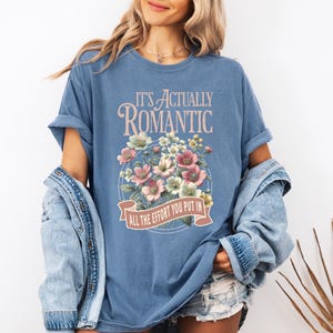 Pu&ograve; includere: Una maglietta blu con la scritta "IT'S ACTUALLY ROMANTIC" sopra un disegno floreale. Il disegno floreale comprende fiori rosa, bianchi e verdi. Il testo "ALL THE EFFORT YOU PUT IN" &egrave; su uno striscione sottostante.
