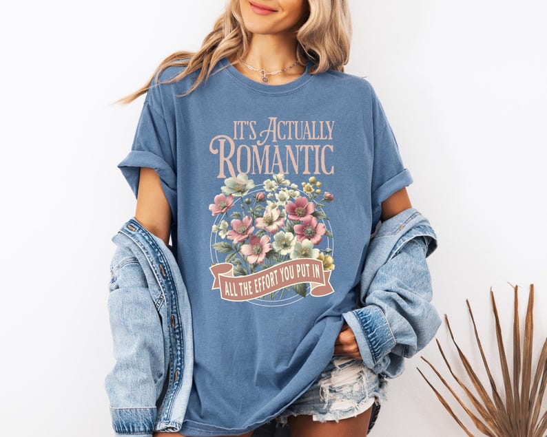 Pu&ograve; includere: Una maglietta blu con la scritta "IT'S ACTUALLY ROMANTIC" sopra un disegno floreale. Il disegno floreale comprende fiori rosa, bianchi e verdi. Il testo "ALL THE EFFORT YOU PUT IN" &egrave; su uno striscione sottostante.