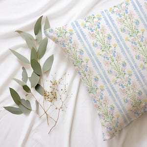 May include: A white pillow with a blue and pink floral pattern. The pillow is on a white bedspread with green leaves and small white flowers.