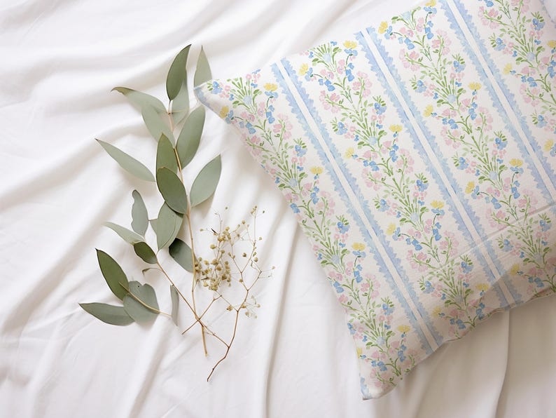 May include: A white pillow with a blue and pink floral pattern. The pillow is on a white bedspread with green leaves and small white flowers.