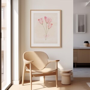 May include: A framed watercolour painting of three pink tulips with a pink ribbon tied around the stems. The painting is hanging on a white wall in a room with a light wood chair and a small table.