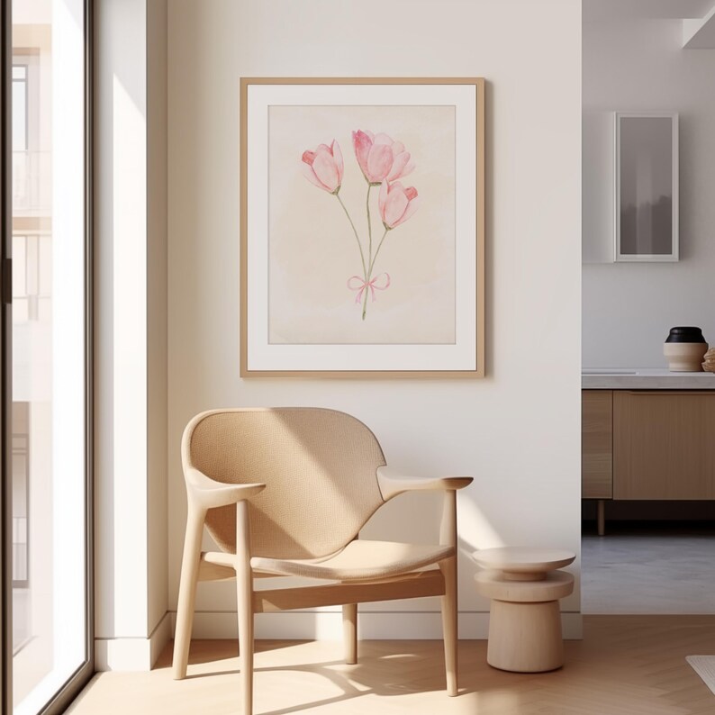 May include: A framed watercolour painting of three pink tulips with a pink ribbon tied around the stems. The painting is hanging on a white wall in a room with a light wood chair and a small table.