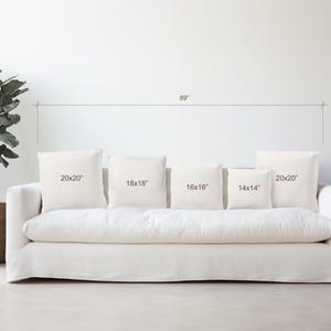 May include: A white sofa with five white throw pillows of different sizes. The pillows are labeled with their dimensions: 20x20 inches, 18x18 inches, 16x16 inches, 14x14 inches, and 20x20 inches.