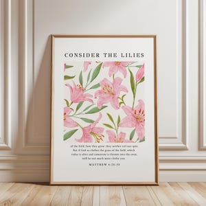 May include: A framed print featuring a floral design of pink lilies and green leaves on a cream background. The text "CONSIDER THE LILIES" is at the top, with a Bible verse below. The frame is a light brown color.