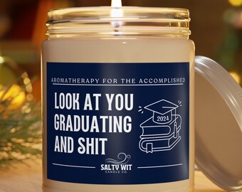 May include: A white candle jar with a blue label that reads "Aromatherapy for the accomplished. Look at you graduating and shit. Salty Wit Candle Co." The label also features a graduation cap and books. The candle jar has a silver lid.