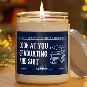 May include: A white candle jar with a blue label that reads "Aromatherapy for the accomplished. Look at you graduating and shit. Salty Wit Candle Co." The label also features a graduation cap and books. The candle jar has a silver lid.