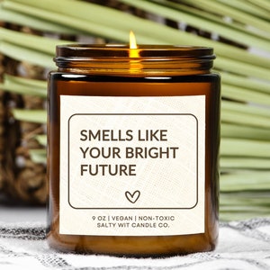 May include: A lit brown glass candle jar with a white label that reads "SMELLS LIKE YOUR BRIGHT FUTURE" with a heart symbol. The candle is 9 oz, vegan, and non-toxic. The background includes a woven basket and green leaves.