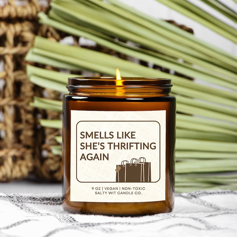 May include: A brown glass candle with a white label that reads "Smells Like She's Thrifting Again". The label also includes a small illustration of a shopping bag and the text "9 oz | Vegan | Non-Toxic Salty Wit Candle Co."