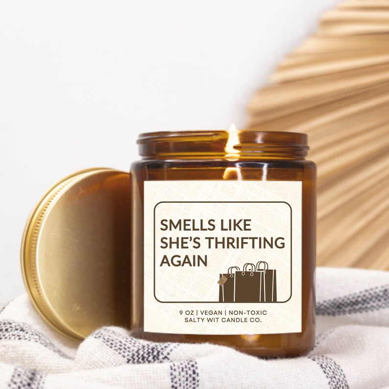 May include: A brown glass candle jar with a white label that reads "Smells like she's thrifting again". The label also includes a graphic of shopping bags and the text "9 oz | Vegan | Non-toxic Salty Wit Candle Co."
