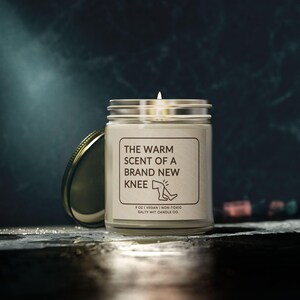 May include: A white candle jar with a gold lid. The label reads "The Warm Scent of a Brand New Knee" with a simple illustration of a knee. The bottom of the label reads "9 oz | Vegan | Non-Toxic Salty Wit Candle Co."