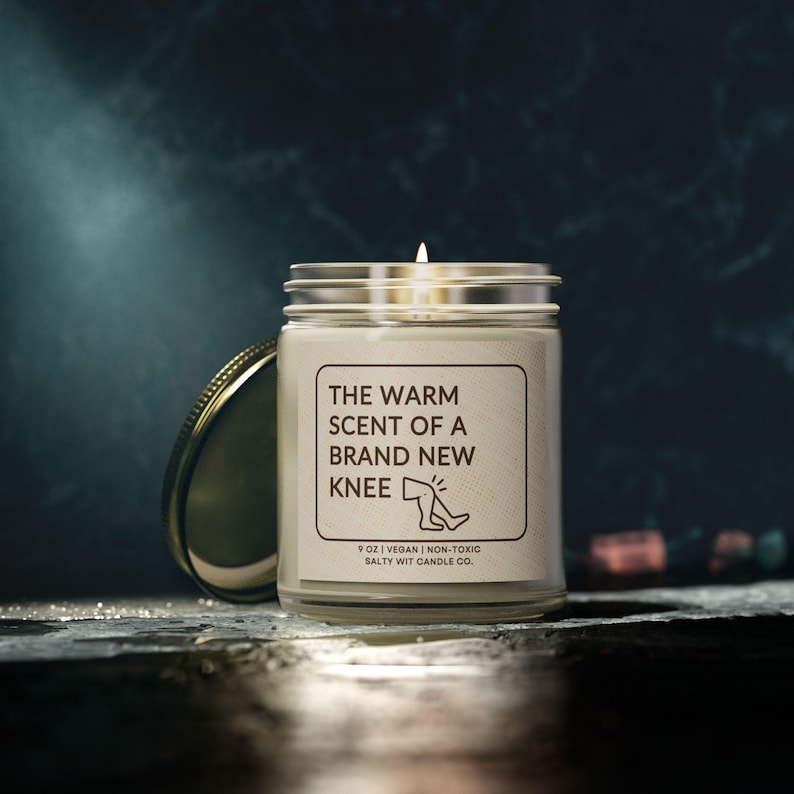 May include: A white candle jar with a gold lid. The label reads "The Warm Scent of a Brand New Knee" with a simple illustration of a knee. The bottom of the label reads "9 oz | Vegan | Non-Toxic Salty Wit Candle Co."