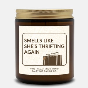 May include: A brown glass candle jar with a gold lid. The label reads "Smells Like She's Thrifting Again" with a graphic of shopping bags. The candle is 9 oz, vegan, and non-toxic. The label also includes the text "Salty Wit Candle Co."