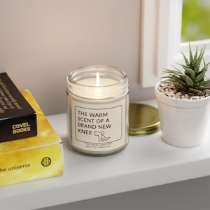 May include: A white candle in a glass jar with a label that reads "The warm scent of a brand new knee". The candle is lit and the label also includes the text "8 oz | Vegan | Non-Toxic | Salty Wit Candle Co."