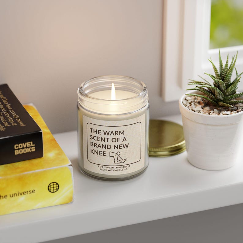 May include: A white candle in a glass jar with a label that reads "The warm scent of a brand new knee". The candle is lit and the label also includes the text "8 oz | Vegan | Non-Toxic | Salty Wit Candle Co."